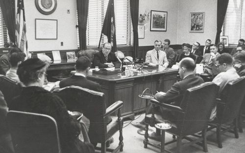 President Benson sitting behind his desk and speaking to a large group of men and a few women.
