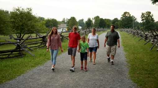 An exterior view of Joseph Smith's family home in Palmyra, New York. 
A family walks by the white building family farm. They are on paths around the farmland.