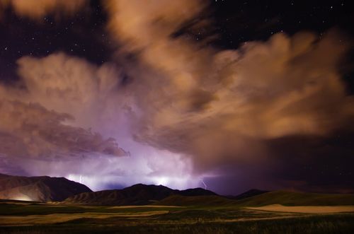 Lightning coming out of big storm clouds at night, striking down over mountains. Stars are seen overhead, above the storm.