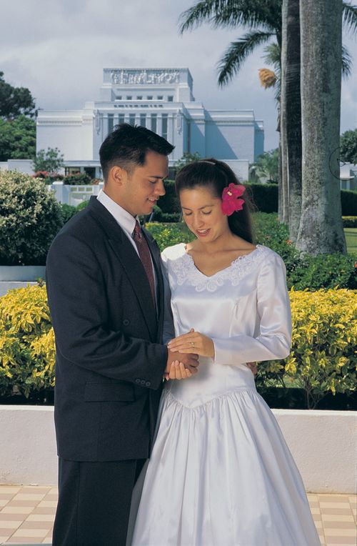 A bride and groom standing in front of the Hawaii Temple.  The couple are holding hands.