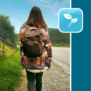 A woman is walking along a road in the mountains. She is wearing a backpack. It appears to be a cloudy or rainy day.