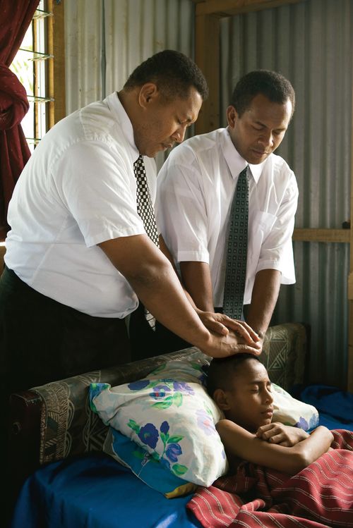 Two Fijian men administering to a young girl lying in a bed.