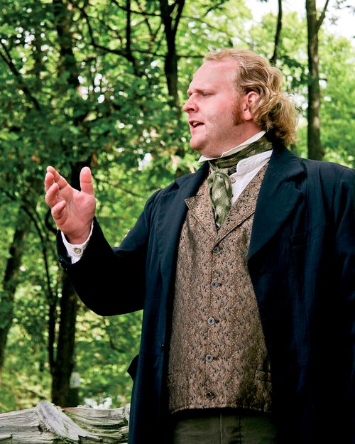 A preacher in the Nauvoo Pageant wearing a green tie, brown vest, and black suit coat, standing and preaching near a group of trees.