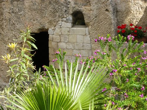 The entrance to the Garden Tomb in Jerusalem, seen beyond large plant fronds and purple flowers growing on a bush.