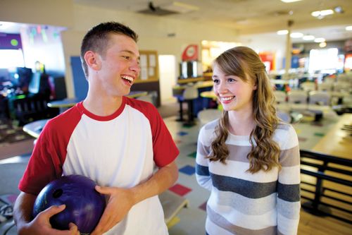A teenage girl at a bowling alley, standing next to a teenage boy, who is laughing and holding a blue bowling ball.