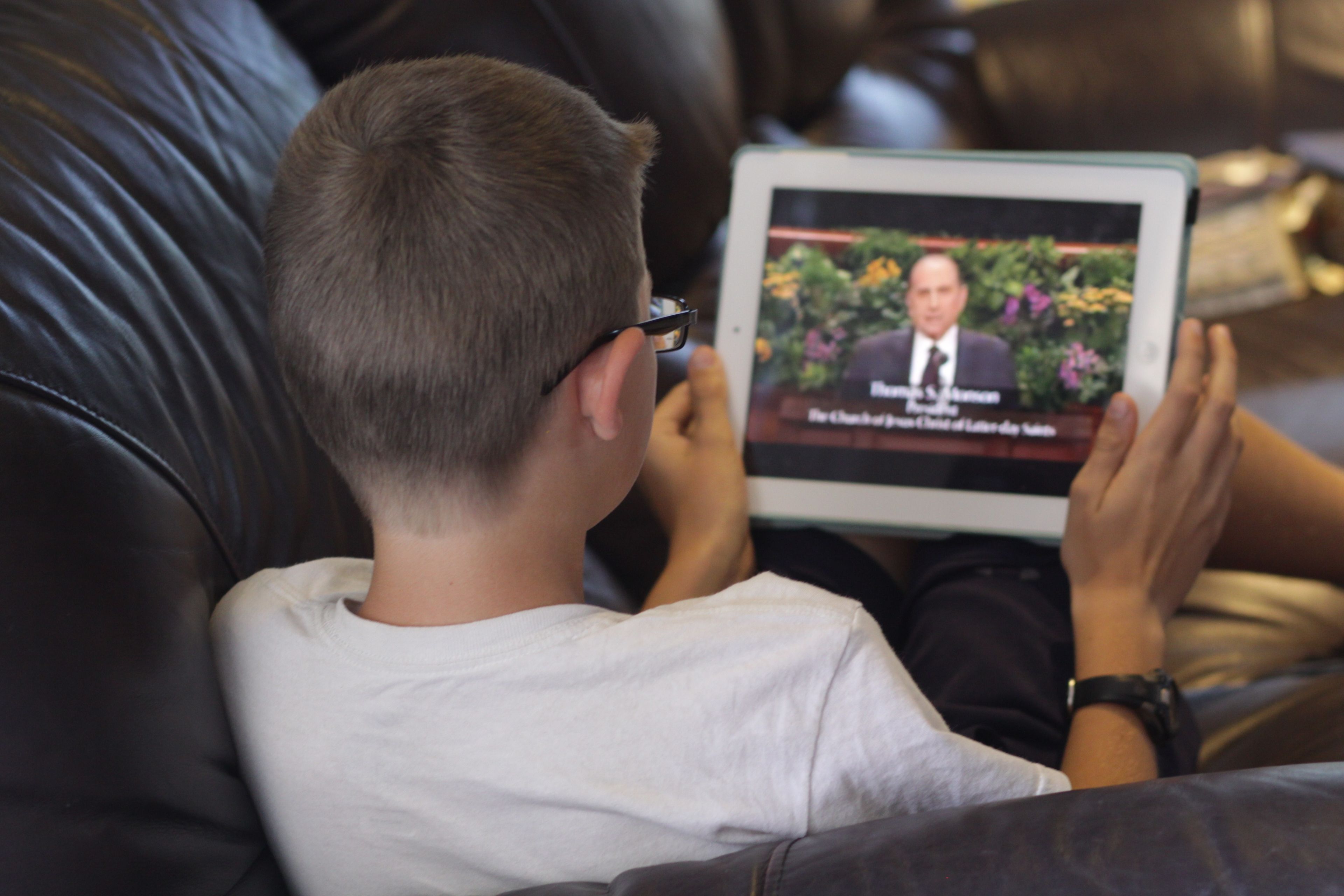 A boy sitting at home watching general conference on his tablet.