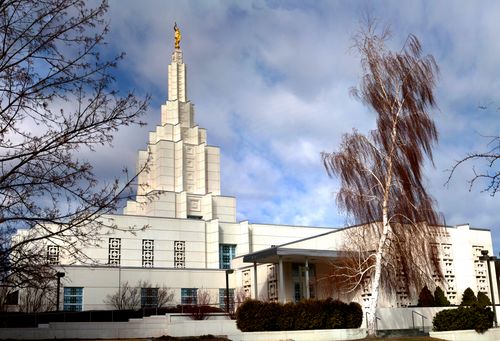 A front side view of the Idaho Falls Idaho Temple on an autumn day, with a tall red tree blowing in the wind.