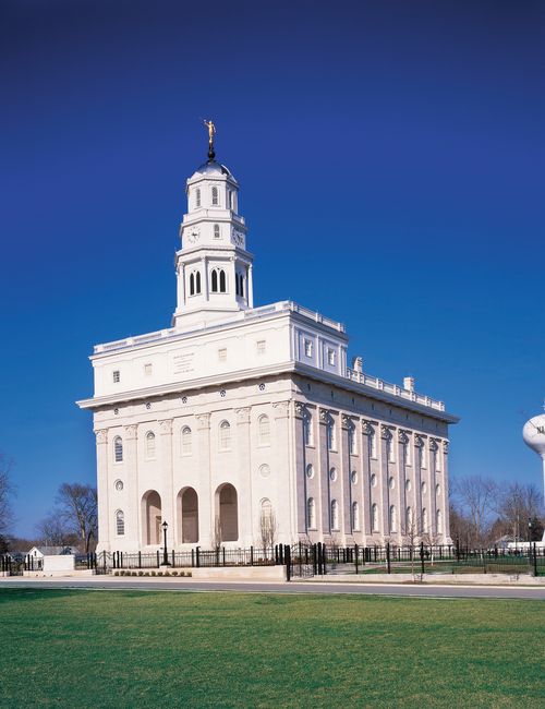The Nauvoo Illinois Temple on a clear, sunny day, with green lawns and a black gate in the foreground.