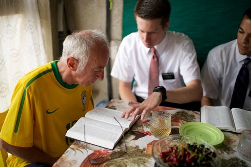 A man in a yellow jersey reads the scriptures along with two elders at his dinner table.