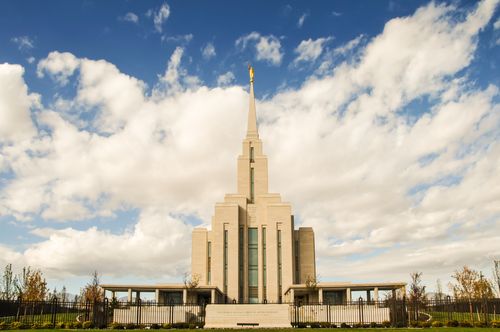 The front of the Oquirrh Mountain Utah Temple on a sunny day, with the temple sign and the black fence seen in the front and large white clouds overhead.