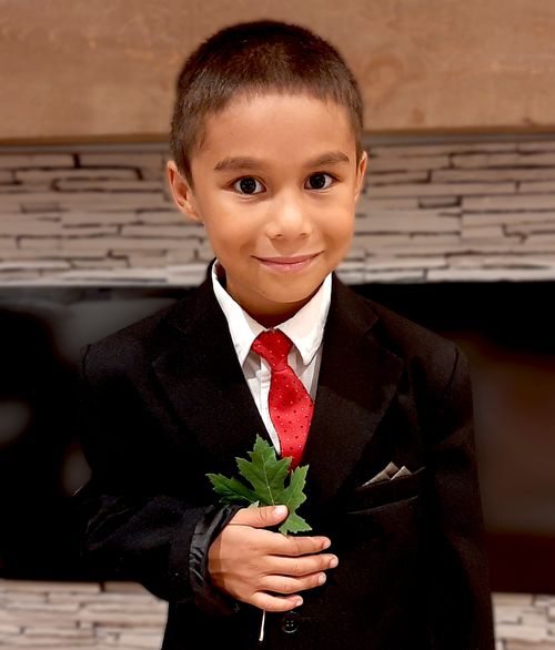 Tristan is dressed in a dark suit with a white shirt and red tie in front of a  a TV. He is holding a leaf.