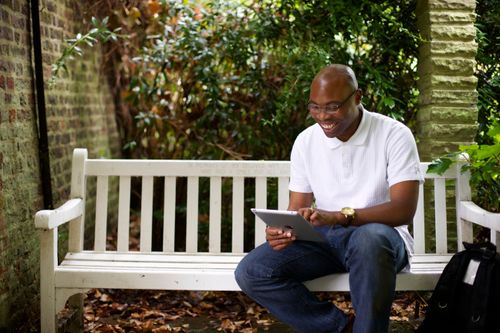 A man in a white shirt, jeans, and glasses sits outside on a white bench and looks at family history on his tablet.