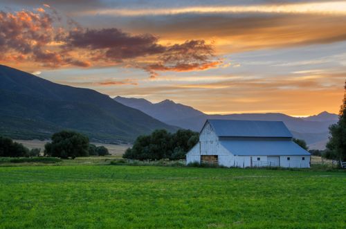 A sunset over a mountain near farmland with a green field, trees, and a large white barn.