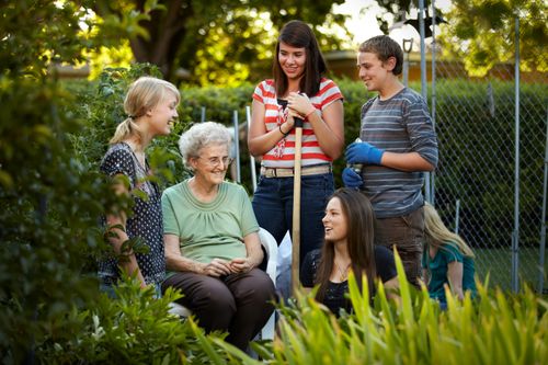 An elderly woman sits on a bench while three young women and one young man help with her garden.