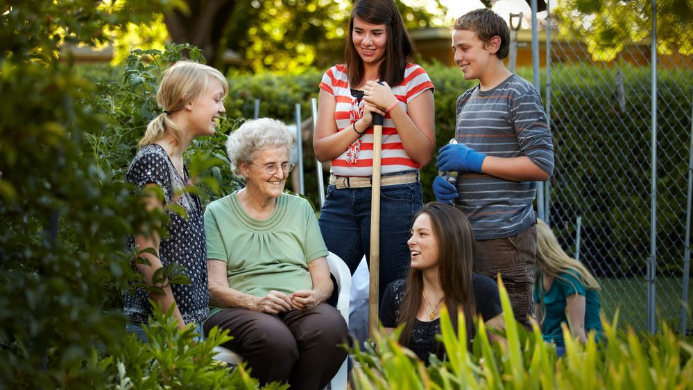 An elderly woman sits on a bench while three young women and one young man help with her garden.