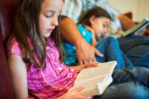 A young girl reads from her scriptures while her father and sister sit next to her on the couch.