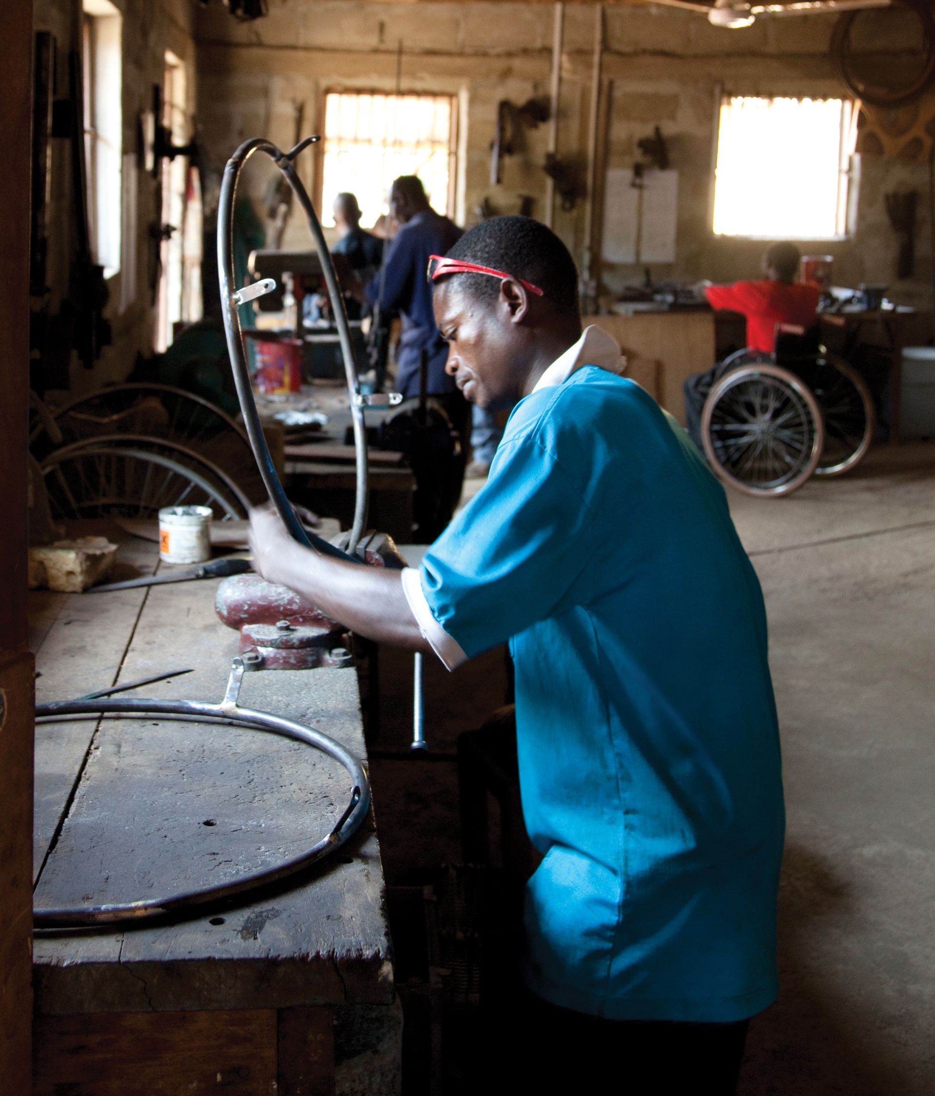 An African man standing in a workshop and repairing a round black wheelchair part with other men nearby.