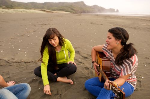 A group of young women sit together on the beach while one plays the guitar.