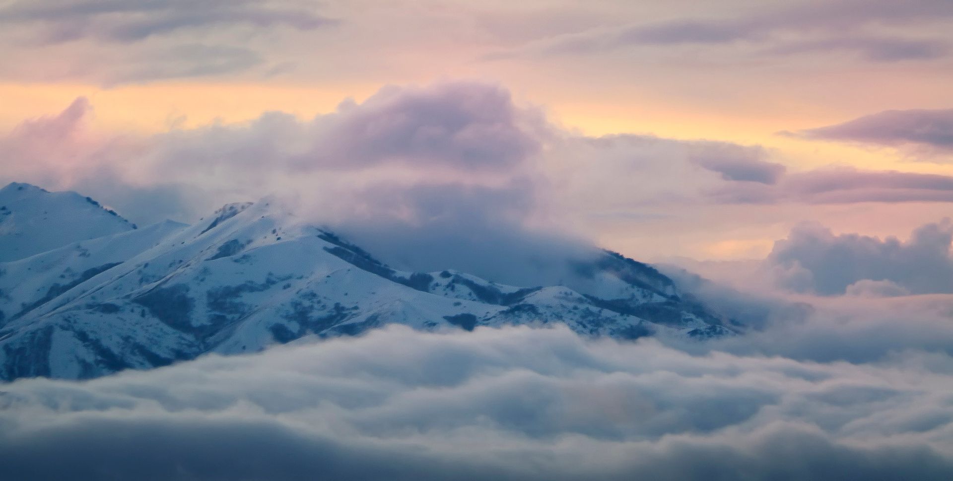 A snow-covered mountain pokes up above a layer of soft clouds as the sun sets.