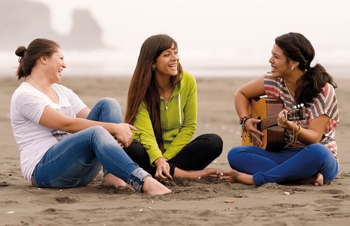 Three young women sitting together on a beach.  One is playing a guitar.