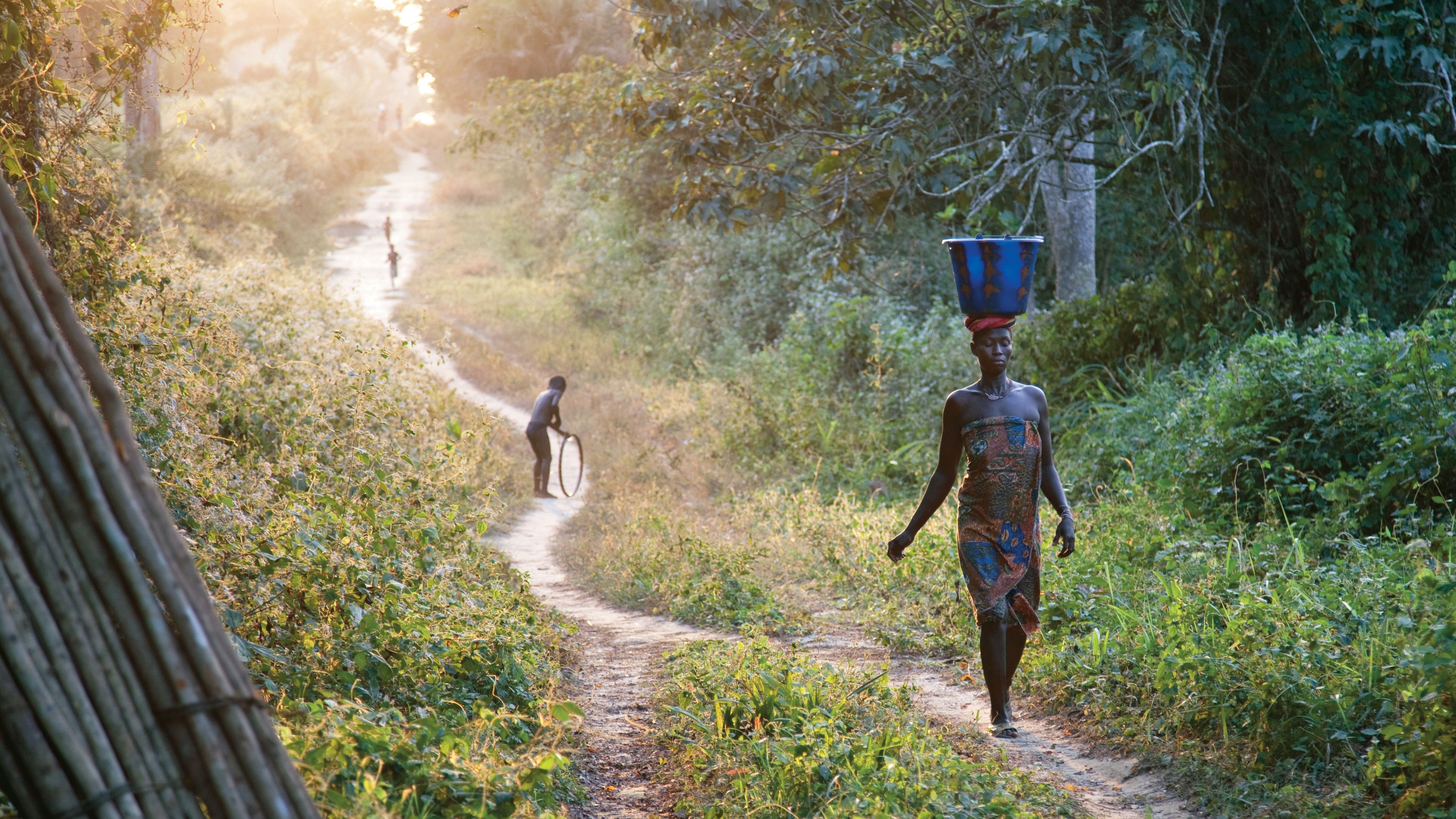 An African woman balancing a bucket on her head while walking down a path.