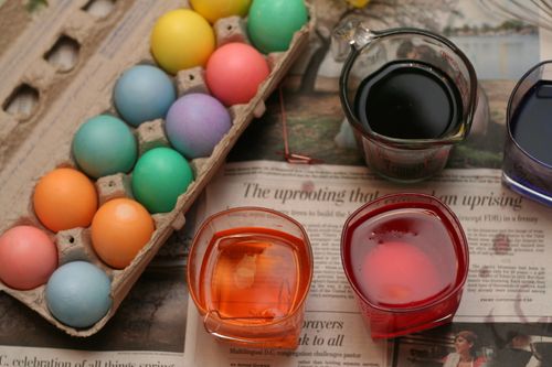 Four cups full of colorful dye sitting on top of a newspaper next to a dozen pastel-colored eggs in a cardboard carton.