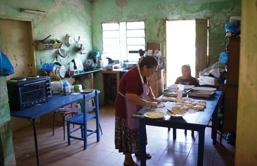 한 청녀가 테이블 옆에 앉아 있고, 한 여성이 테이블에서 빵을 만들고 있다.