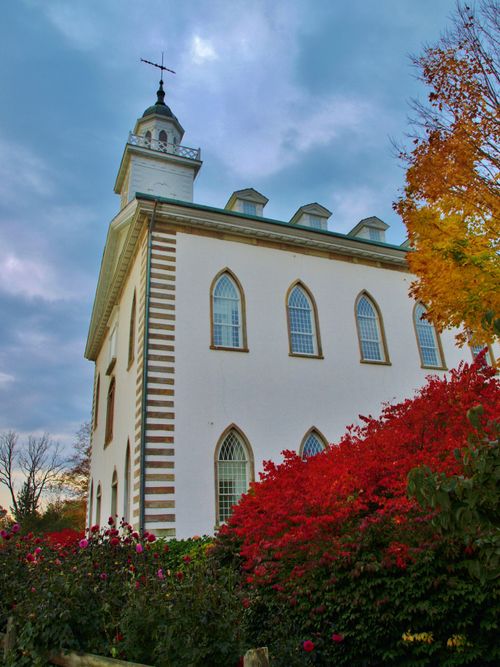 A view of the Kirtland Temple from below, looking upward on a fall day with a red bush in the foreground.