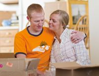 A young couple sitting next to each other in an apartment or house.  There are boxes around them to indicate they just moved in.  They are smiling and hugging each other.