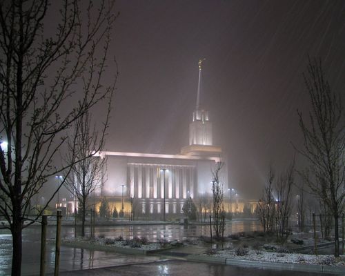 A side view of the Oquirrh Mountain Utah Temple on a snowy evening, with bare trees in the parking lot medians.