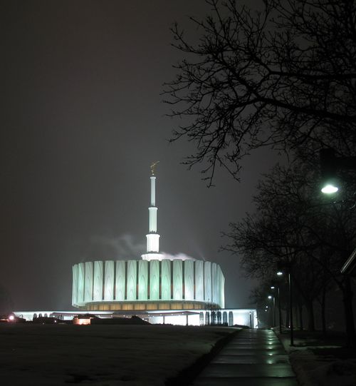 The Provo Utah Temple on a winter night, seen from the bottom of the hill, with the silhouette of a large bare tree on the right side.