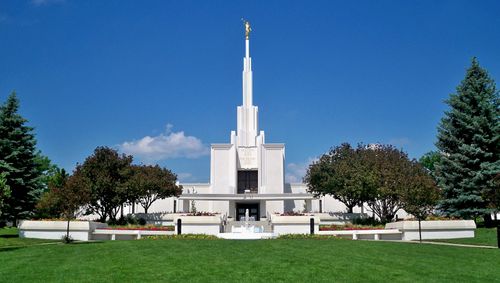 A front view of the Denver Colorado Temple on a sunny day, with the green lawn in front and large trees growing on the grounds.