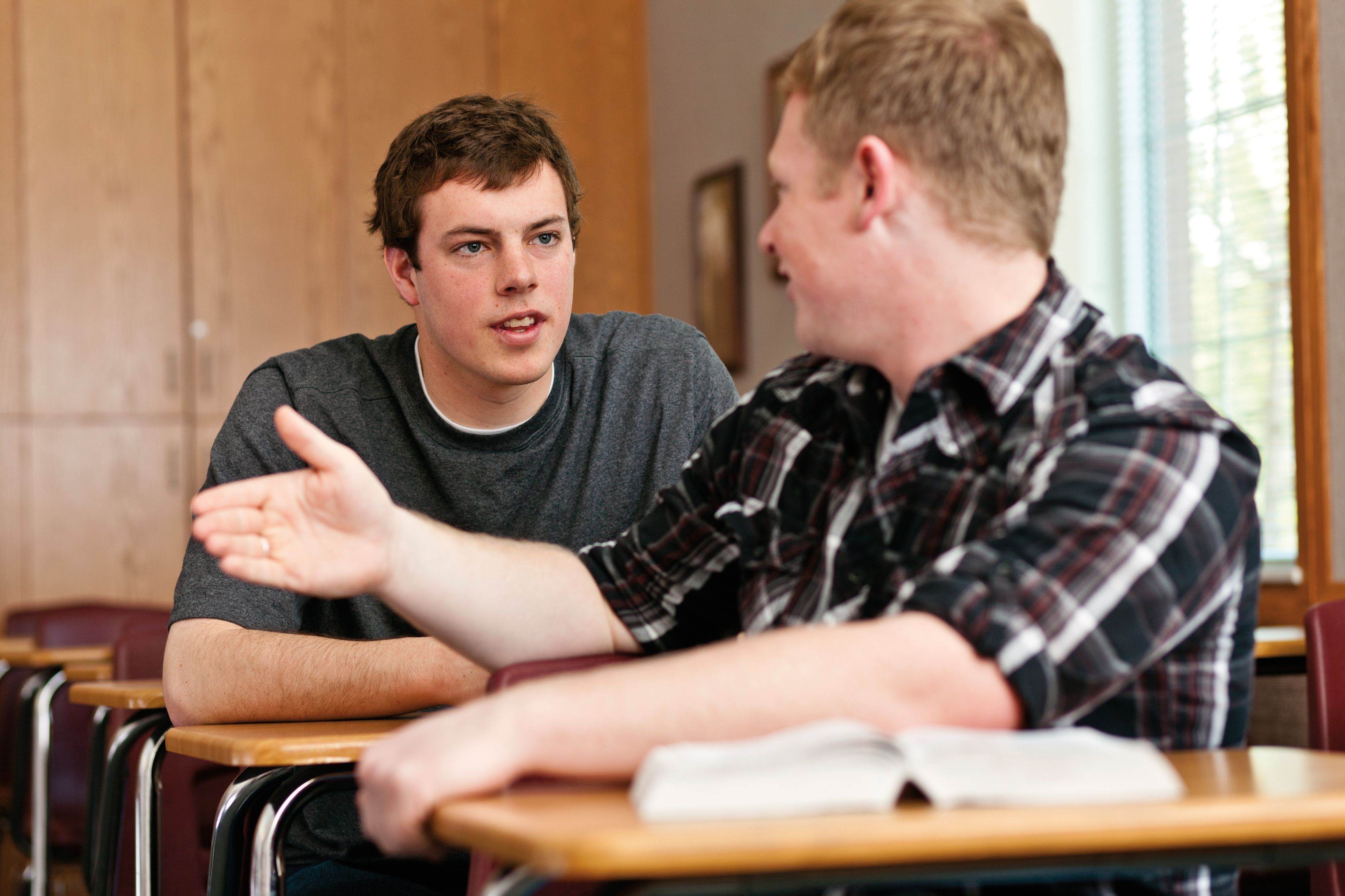 Two young men sit in a classroom together with their scriptures open as they talk.