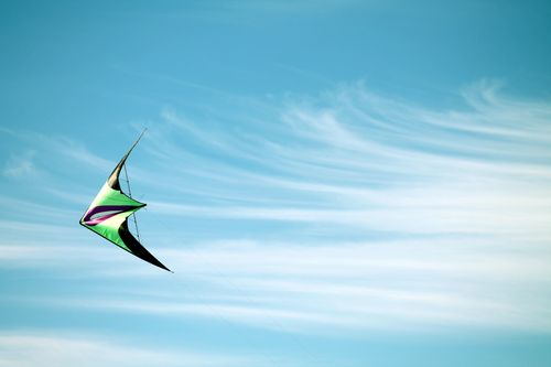 A green, purple, and black kite flies through a cloudy blue sky.