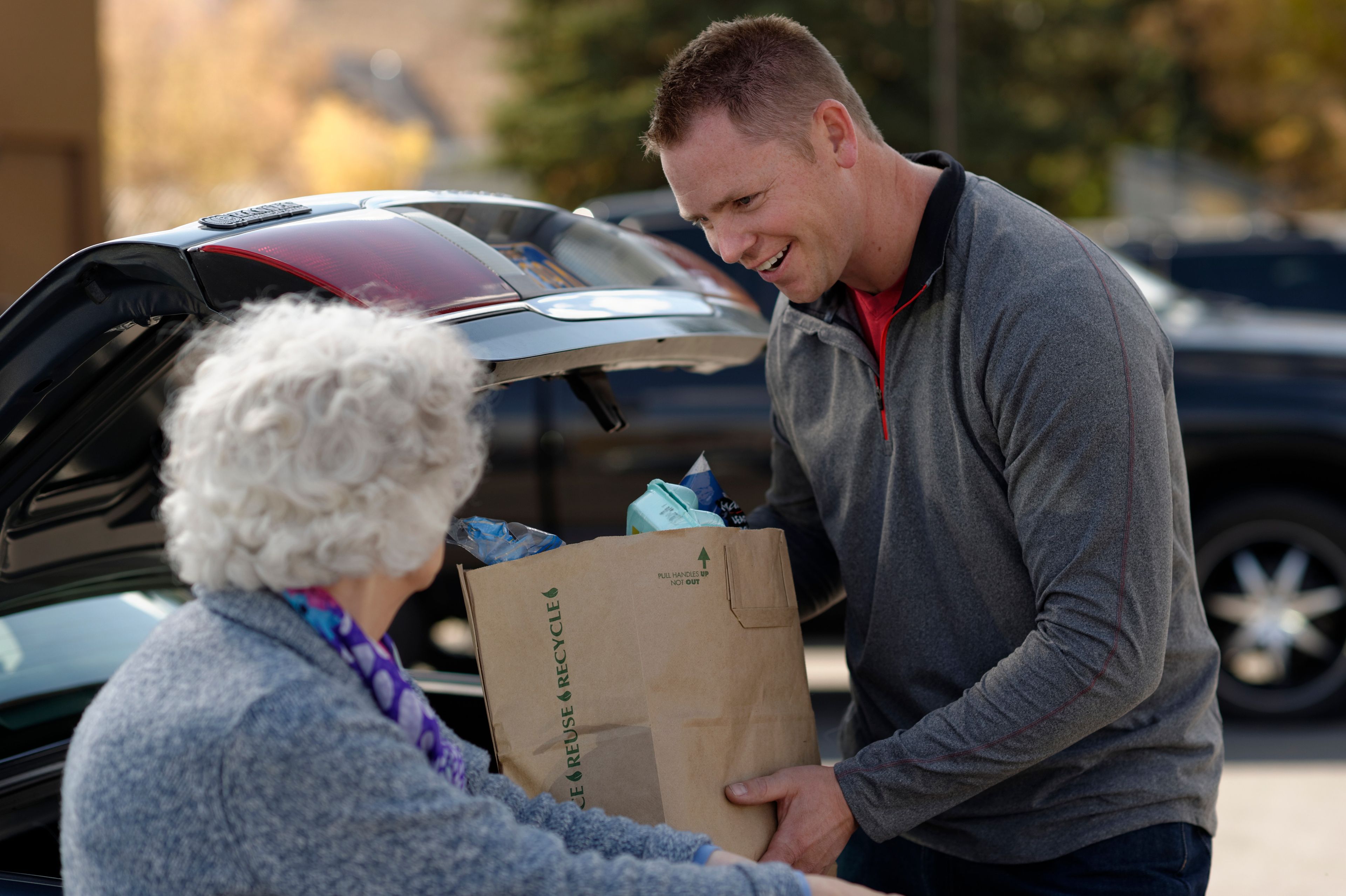 A man helping an elderly woman with her groceries in a parking lot.