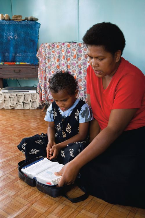 A mother sits on the floor next to her young daughter and reads from the scriptures as her daughter listens.