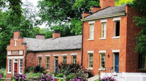 A complex of three connected red brick buildings. There is a sign on the last section that reads: “Jonathan Browning Gunsmith Blacksmith & Farrier.”