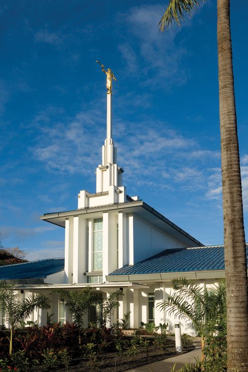 A portrait view of the Papeete Tahiti Temple on a sunny day, with a blue sky overhead and a palm tree on the right side.