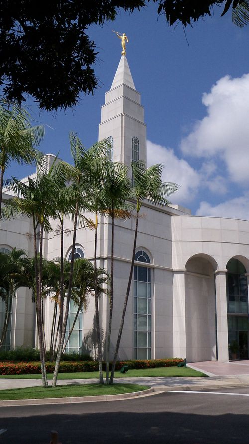 A partial side view of the Recife Brazil Temple through trees on the grounds around the temple, including a view of some arches and the spire.