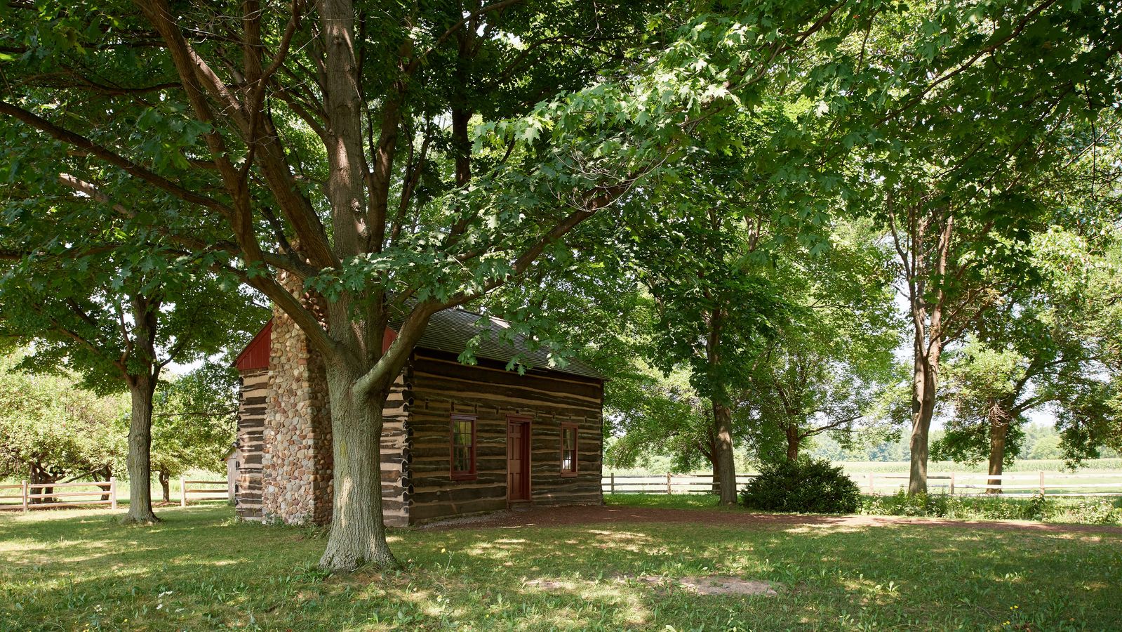 Exterior shots of the Peter and Mary Whitmer Farm. There is a log cabin with a red door. It is surrounded by trees.