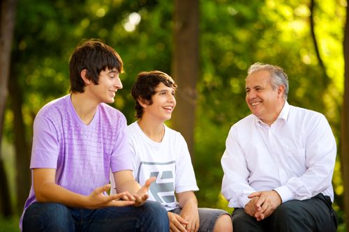 Two young men from Argentina sitting and laughing with an older man outside near trees with green and yellow leaves.