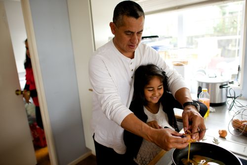 A father cracks an egg with his daughter over a skillet while they cook in the kitchen.