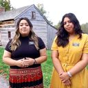 Two sister missionaries standing in front of a historic log cabin.