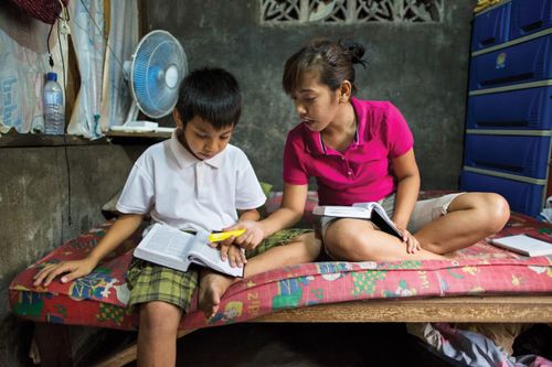 A girl in a pink shirt sitting on a bed next to her brother as they study the scriptures.