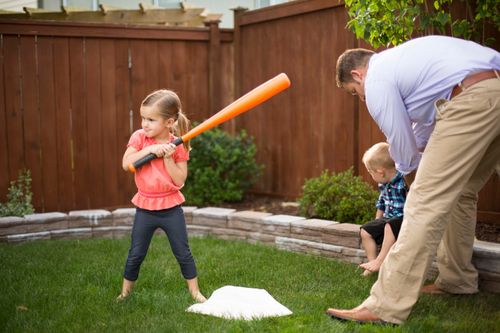 A young girl stands next to a base, holding an orange bat, with her father and younger brother nearby.