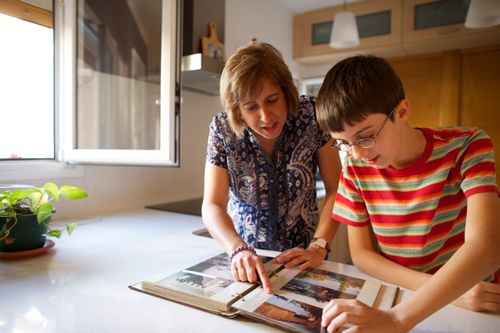 A grandmother looks at family pictures in a photo album with her grandson at the kitchen counter.