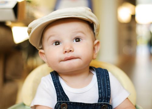 A baby boy in overalls and a hat, sitting and looking up.