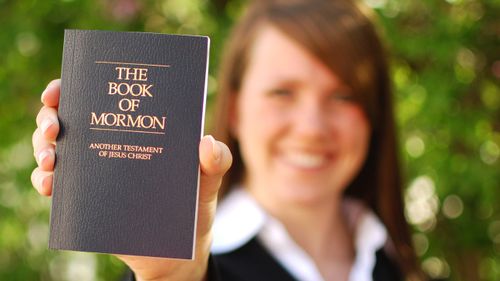 A woman smiles as she holds up the Book of Mormon. The focus is on the Book of Mormon. She is blurred.