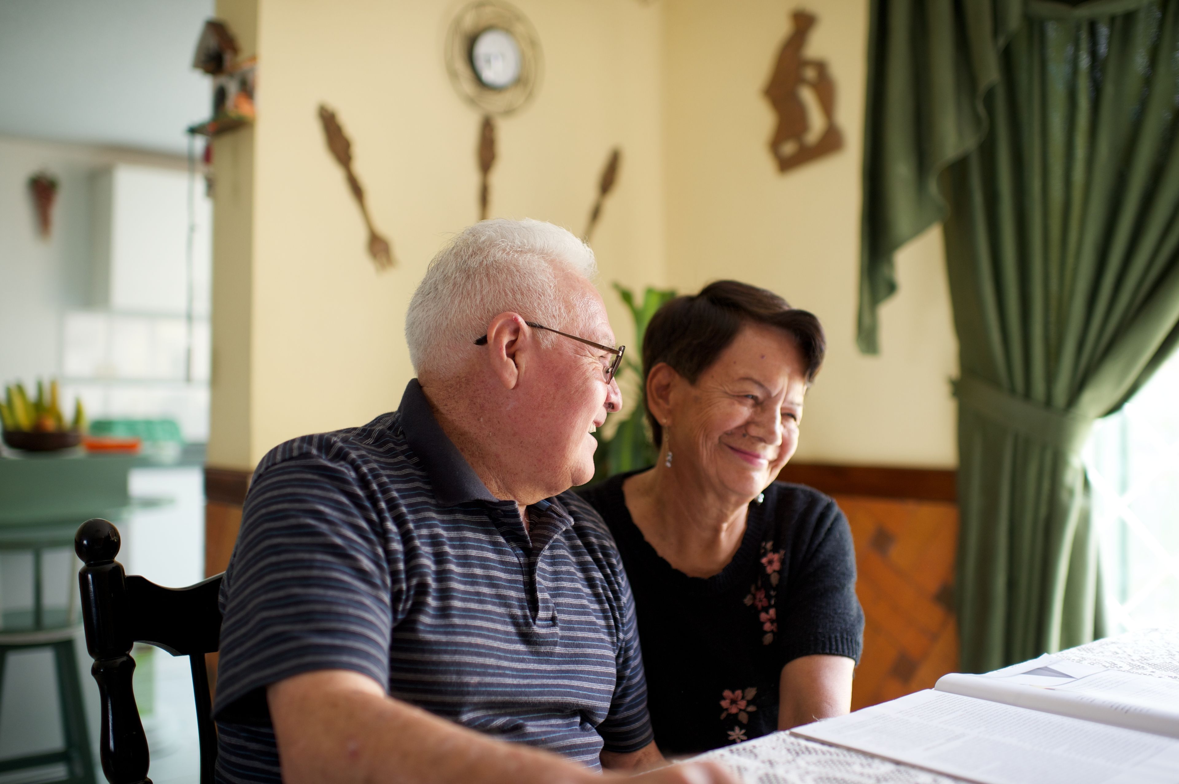 An elderly couple sits in chairs at their house and laughs together.