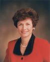 A photograph of Margaret Dyreng Nadauld against a brown background, wearing a red blazer with a black collar.